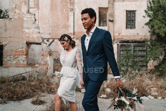 Jeune marié multiracial heureux et mariée tenant la main se promener sur la passerelle contre la construction âgée le jour du mariage — Photo de stock