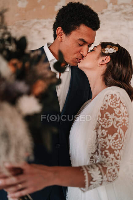 Side view of loving young ethnic newlywed couple wearing chic wedding clothes embracing and kissing gently in obsolete remaining building — Stock Photo