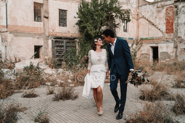 Jovem feliz noivo multirracial e noiva olhando um para o outro enquanto passeia na passarela contra a construção envelhecida no dia do casamento — Fotografia de Stock
