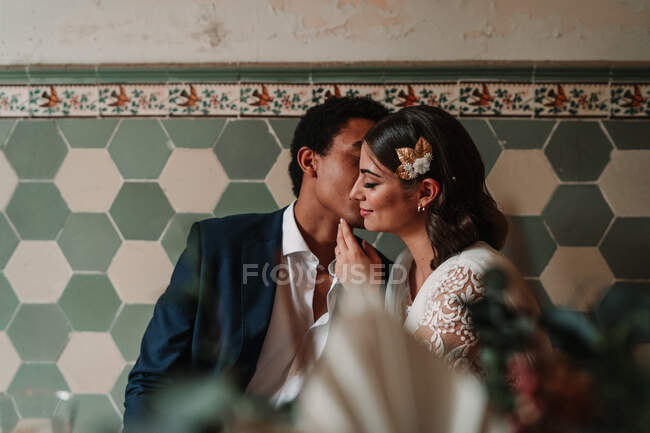 Anonymous ethnic man in suit whispering in ear of content beloved with closed eyes while sitting at restaurant table on wedding day — Stock Photo