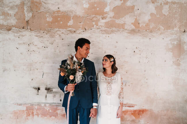 Молодий усміхнений багаторасовий наречений з весільним букетом тримає кохану вручну, дивлячись один на одного біля старої стіни — стокове фото