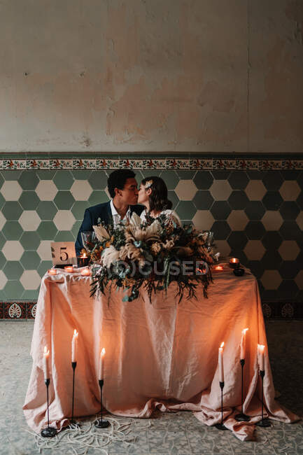 Junge multiethnische Braut und Bräutigam sitzen am Tisch und küssen sich mit Blumenstrauß und brennenden Kerzen mit geschlossenen Augen — Stockfoto