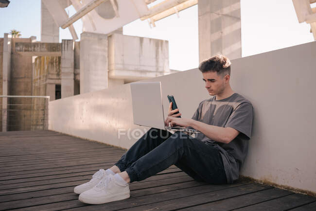 Cuerpo completo de joven freelancer masculino concentrado en traje de moda navegando en el teléfono móvil mientras trabaja de forma remota en el portátil sentado en la terraza en el día soleado - foto de stock