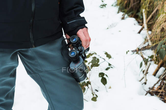 Escursionista maschio irriconoscibile ritagliato in abiti caldi in piedi in boschi invernali innevati con binocolo — Foto stock