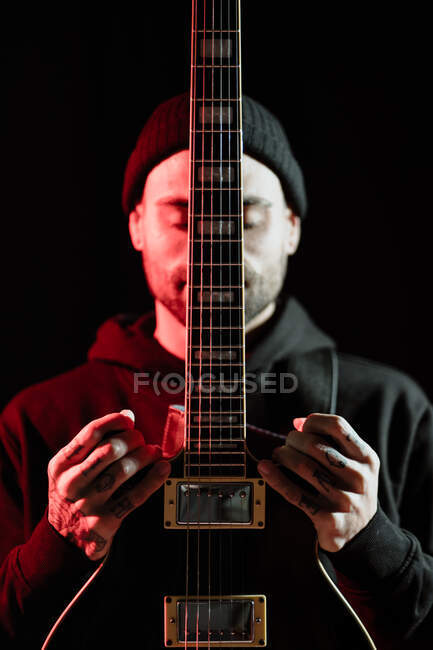 Спокойный рок-музыкант, стоящий с электрогитарой на черном фоне в студии с красным светом — стоковое фото
