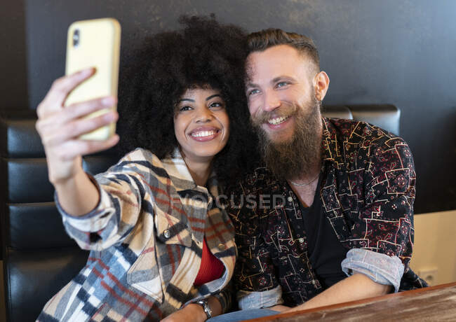 Захоплена багаторасова пара сидить за столом в кафе і приймає селфі на смартфон — стокове фото