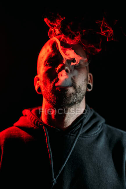 Серйозний чоловічий рокер палить сигарету і дивиться на камеру на чорному тлі в студії з червоним освітленням — стокове фото