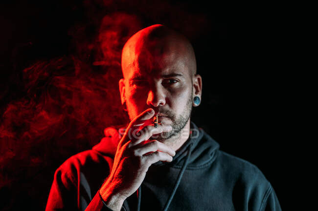 Grave rockero masculino fumando cigarrillo y mirando a la cámara en el fondo negro en el estudio con iluminación roja - foto de stock
