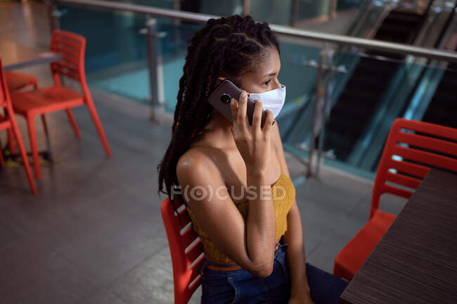 Retrato de una atractiva joven latina afro vestida con una mascarilla y hablando en un smartphone en un centro comercial, Colombia - foto de stock