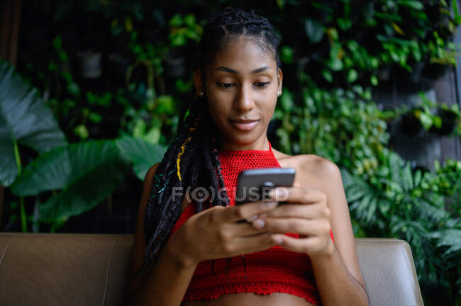 Retrato de atraente jovem afro mulher latina com dreadlocks em um top vermelho de crochê usando smartphone na mesa de restaurante, Colômbia — Fotografia de Stock