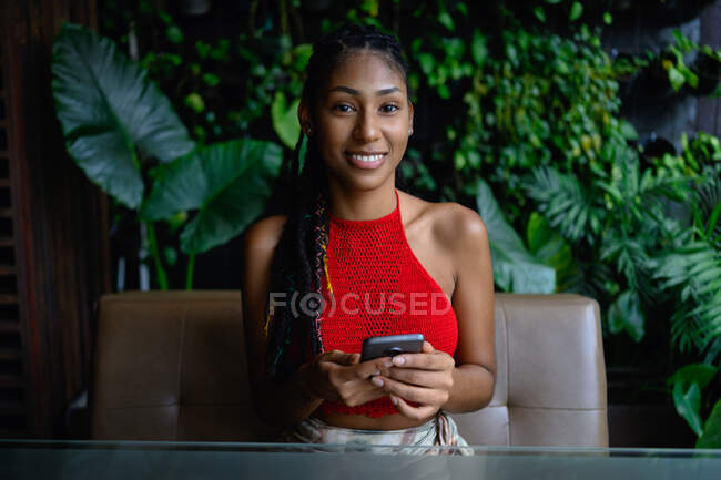 Porträt einer attraktiven jungen Afro-Lateinerin mit Dreadlocks in einem gehäkelten roten Top, die mit Smartphone am Restauranttisch posiert, Kolumbien — Stockfoto