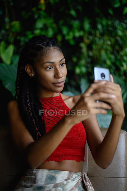 Retrato de atractiva joven afro latina con rastas en un top rojo del ganchillo usando smartphone en mesa de restaurante, Colombia - foto de stock