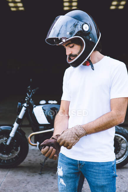 Concentrated young unshaven ethnic male biker in casual outfit and helmet wearing gloves while standing near motorcycle on street — Stock Photo