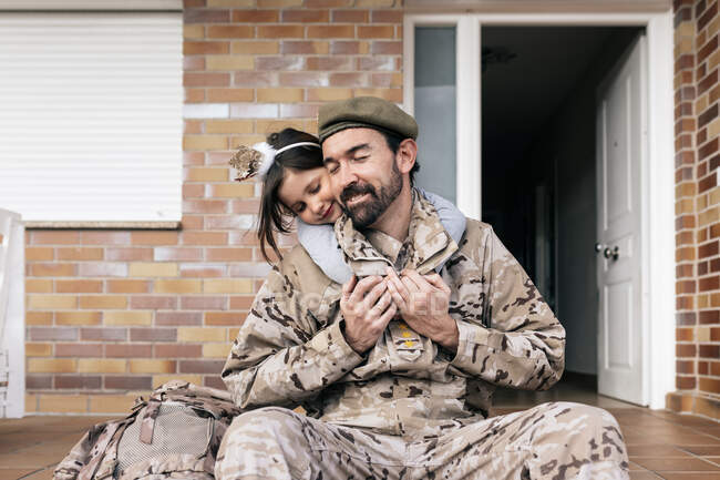 Filha bonito ternamente abraçando pai em uniforme militar sentado à porta após a chegada — Fotografia de Stock