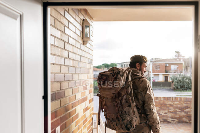 Задний вид мужчины-солдата с рюкзаком и в военной форме, стоящего перед дверью перед отъездом — стоковое фото