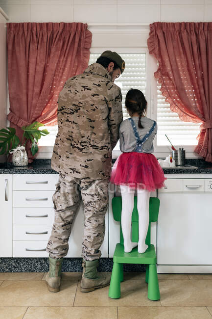 Visão traseira do pai militar de uniforme de pé com criança na cozinha e lavar pratos juntos — Fotografia de Stock