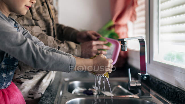 Вид сбоку на военнослужащего в форме, стоящего у раковины с очаровательной дочерью, моющей посуду вместе на кухне дома — стоковое фото