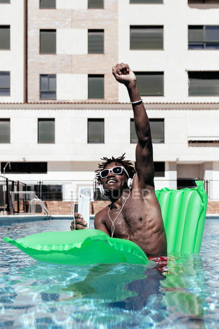 Приємний афроамериканець сидить на надувному матраці в басейні і слухає музику в навушниках під час літніх канікул з піднятою рукою. — Stock Photo