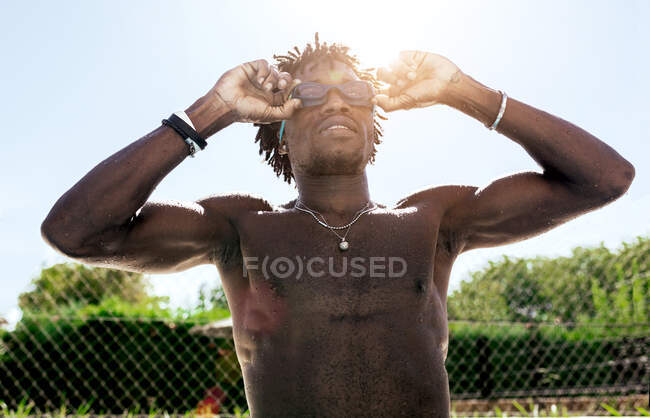 Уверенный молодой афро-американский спортсмен с обнаженным туловищем, регулирующий очки и поднимающий взгляд, стоя у бассейна после купания — стоковое фото