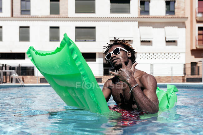 Крутой афроамериканец в солнцезащитных очках, сидящий на надувной матрасе и показывающий рок-жест, глядя в камеру и наслаждаясь выходными в бассейне — стоковое фото