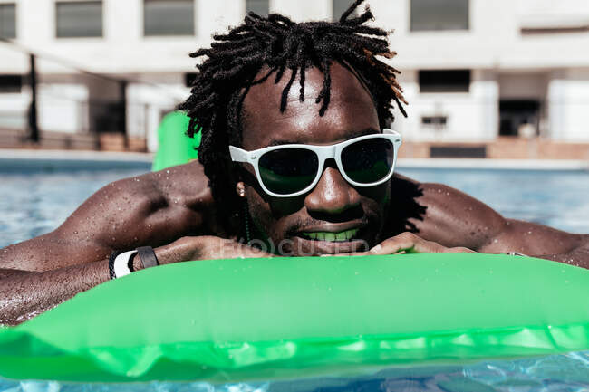 Homem afro-americano fresco em óculos de sol deitado em um colchão inflável e desfrutando do fim de semana na piscina — Fotografia de Stock