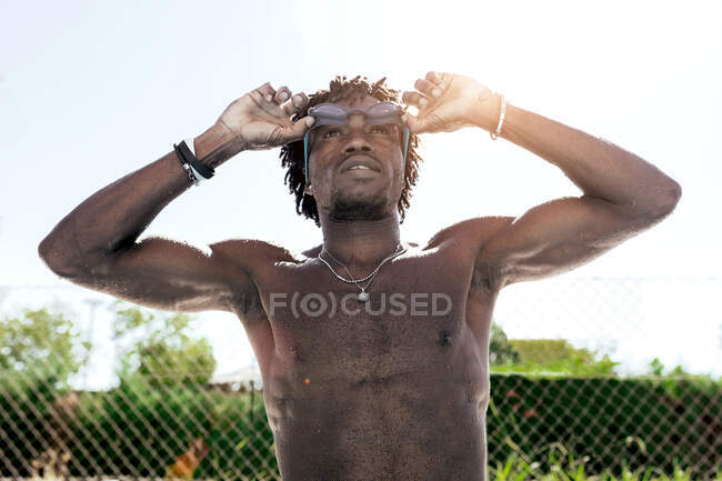Fiducioso giovane atleta afroamericano maschio con busto nudo regolazione occhiali e guardando in alto mentre in piedi a bordo piscina dopo il nuoto — Foto stock