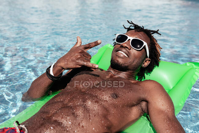 Homem afro-americano legal em óculos de sol deitado em um colchão inflável e mostrando um gesto de rocha enquanto olha para a câmera e desfruta do fim de semana na piscina — Fotografia de Stock