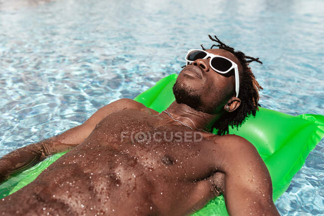 Homem afro-americano fresco em óculos de sol deitado em um colchão inflável e desfrutando do fim de semana na piscina — Fotografia de Stock