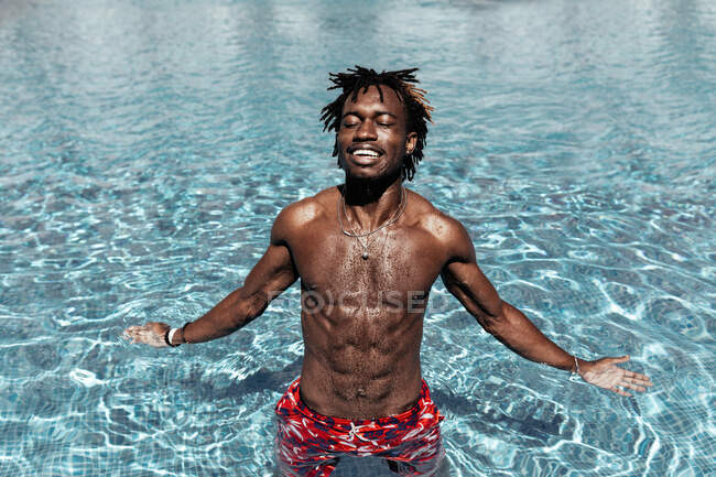Homme afro-américain insouciant debout avec les yeux fermés dans la piscine et profiter des vacances d'été — Photo de stock