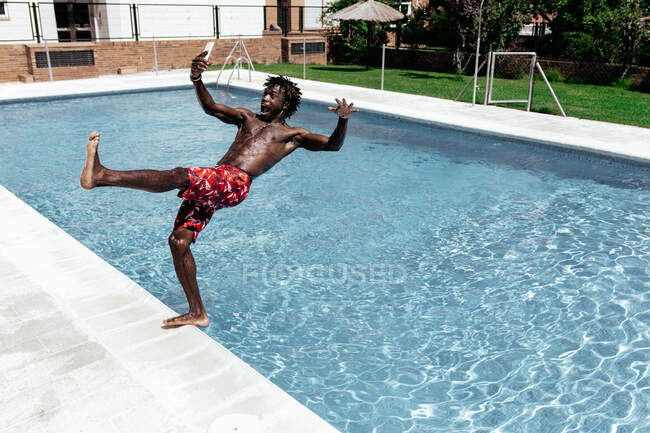 Side view of astonished African American male in shorts and with mobile phone falling into swimming pool — Stock Photo