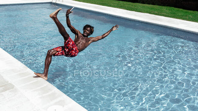 Astonished African American male in shorts and with mobile phone falling into swimming pool — Stock Photo