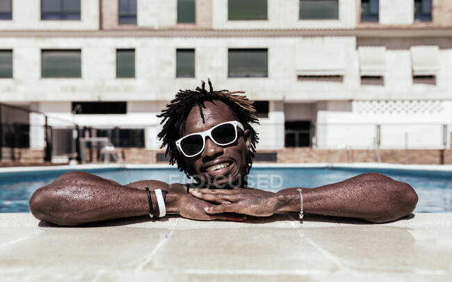 Entzückter Afroamerikaner mit Sonnenbrille lehnt am Pool, genießt den Sommerurlaub und blickt in die Kamera — Stockfoto