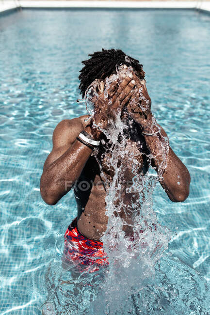 Anonymer Afroamerikaner in kurzen Hosen steht im Schwimmbad und spritzt Wasser ins Gesicht, während er das Sommerwochenende an einem sonnigen Tag genießt — Stockfoto