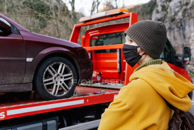 Side view emotionless female in outerwear and protective mask standing near tow truck with car after breakdown in autumn countryside — Stock Photo