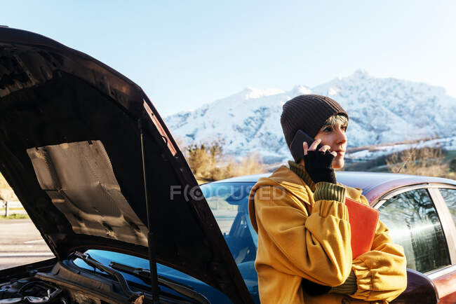 Calm young female in warm clothes having phone call and leaning on car with opened hood while standing on roadside and looking away after breakdown on sunny autumn day — Stock Photo