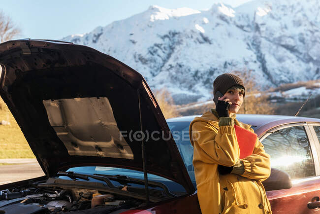 Calm young female in warm clothes having phone call and leaning on car with opened hood while standing on roadside and looking away after breakdown on sunny autumn day — Stock Photo