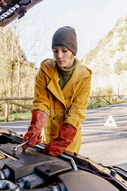 Mujer joven en ropa de abrigo y guantes no conductores aplicando abrazaderas en la batería del coche bajo el capó en el soleado parque de otoño - foto de stock