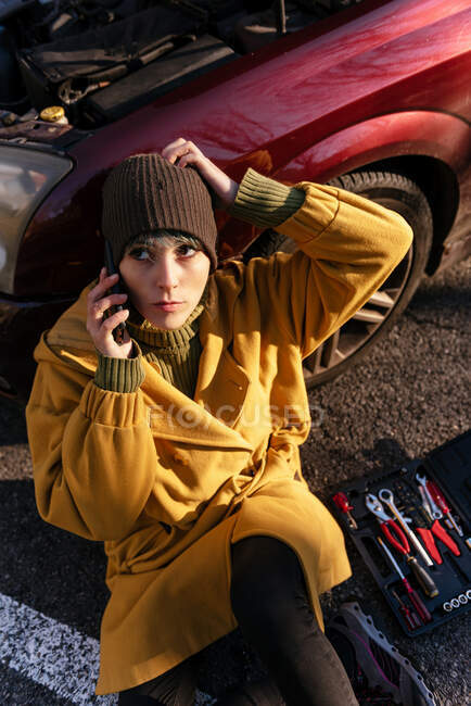 Calm young female in warm clothes having phone call and sitting on the asphalt leaning on car with opened hood near tool box on roadside and looking away after breakdown on sunny autumn day — Stock Photo