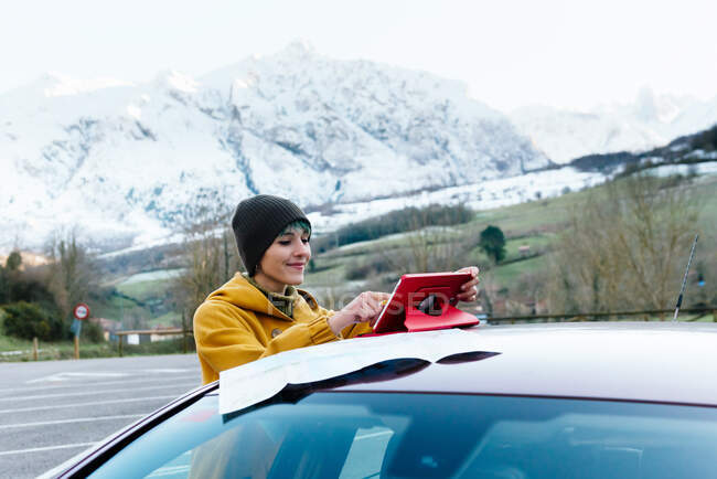 Contenuto giovane donna in abiti caldi navigazione tablet sul tetto dell'auto mentre in piedi su un terreno montagnoso il gelo giorno d'inverno — Foto stock