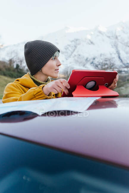 Вміст молодої жінки в теплому одязі, що переглядає планшет на даху автомобіля і дивиться геть, стоячи на гірській місцевості в морозний зимовий день — стокове фото