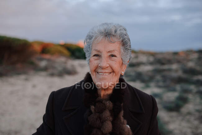 Feliz turista anciana con el pelo gris en traje casual cálido sonriendo y mirando a la cámara mientras se relaja en la playa de arena contra el cielo nublado de la noche - foto de stock