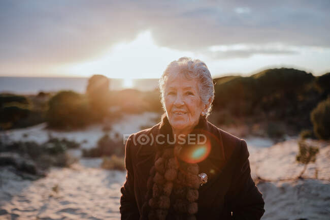 Heureux touriste âgée avec des cheveux gris dans une tenue décontractée chaude souriant et regardant la caméra tout en se relaxant sur la plage de sable contre ciel nuageux en soirée — Photo de stock