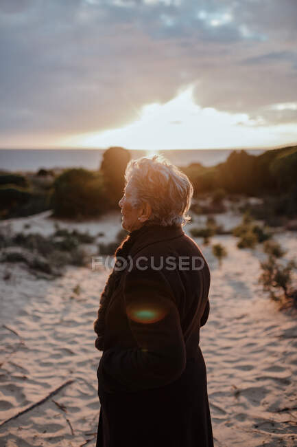 Elderly female tourist with gray hair in warm casual looking away while relaxing on sandy beach against cloudy evening sky — Stock Photo
