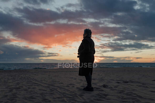 Вид сбоку на неузнаваемую пожилую женщину в теплой повседневной одежде, любующуюся закатом над морем, отдыхающую одну на песчаном пляже — стоковое фото