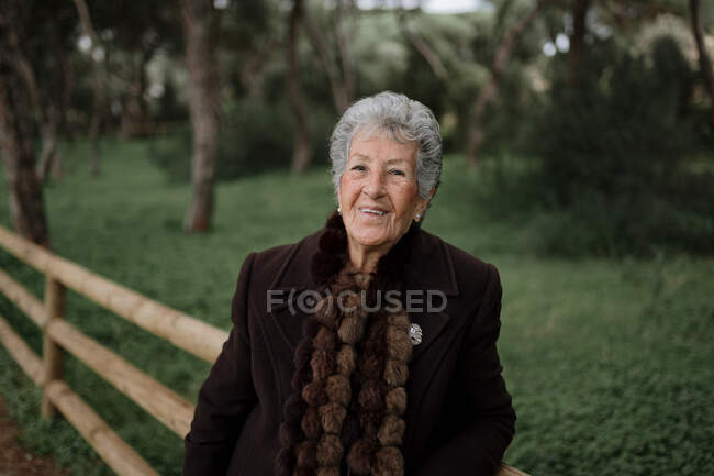 Пожилая женщина-путешественница в повседневной одежде стоит на деревянном пирсе на природе рядом с пляжем и наслаждается морским пейзажем и смотрит в камеру — стоковое фото