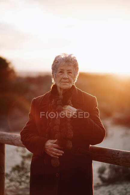 Viajera anciana vestida con ropa casual parada en un muelle de madera en la playa de arena y disfrutando del paisaje marino al atardecer - foto de stock