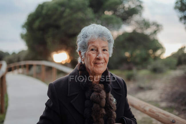 Viandante anziana donna in abiti casual in piedi su un molo di legno sulla spiaggia di sabbia e godendo paesaggio marino e guardando la fotocamera — Foto stock