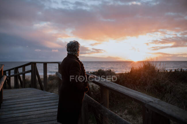 Боковой вид на анонимную пожилую женщину-путешественницу в повседневной одежде, стоящую на деревянном пирсе на песчаном пляже и наслаждающуюся морским пейзажем на закате — стоковое фото