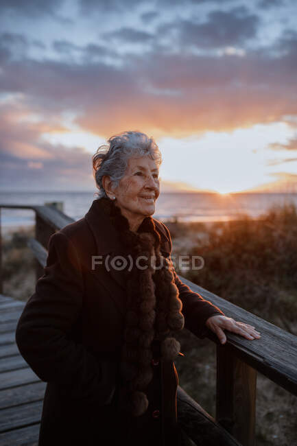 Vista lateral de una anciana viajera vestida con ropa casual parada en un muelle de madera en la playa de arena y disfrutando del paisaje marino al atardecer - foto de stock