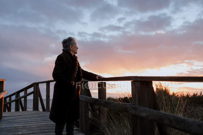 Вид на пожилую женщину-путешественницу в повседневной одежде, стоящую на деревянном пирсе на песчаном пляже и наслаждающуюся морским пейзажем на закате — стоковое фото